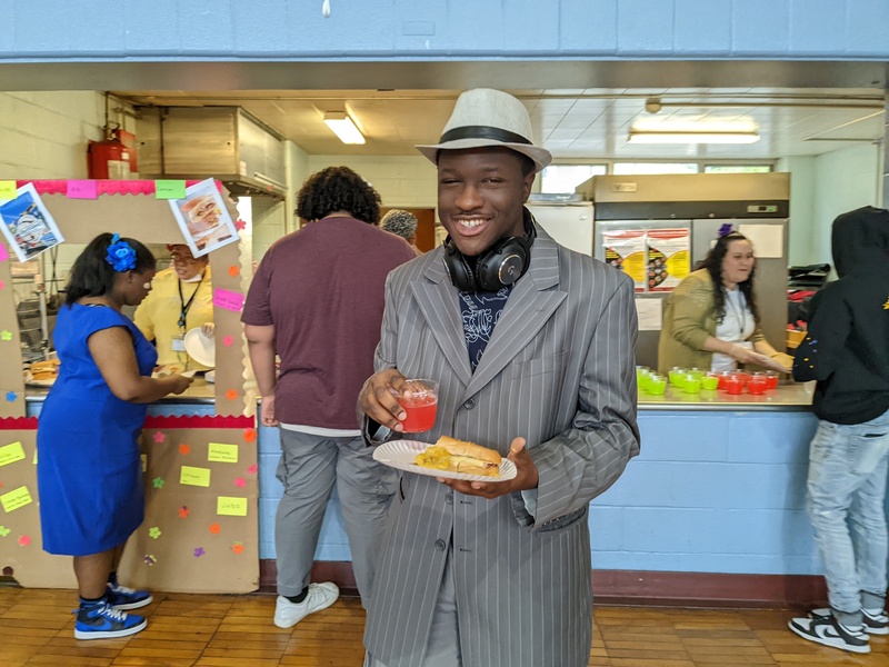 a student dressed in a suit and tie holds a plate of food and smiles with the kitchen in the background serving students